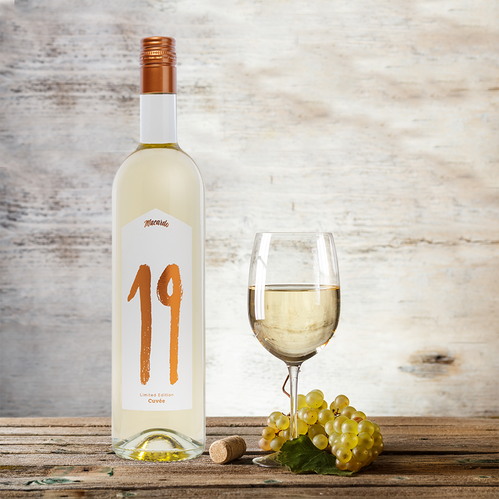 Eine Flasche Weisswein "19" Cuvée steht neben einem Weinglas auf einem Holztisch. Vor dem Glas liegen grüne Trauben und ein Korken. Die Flasche hat ein weißes Etikett mit der goldenen Aufschrift "19". Der Hintergrund ist eine helle Holzwand.